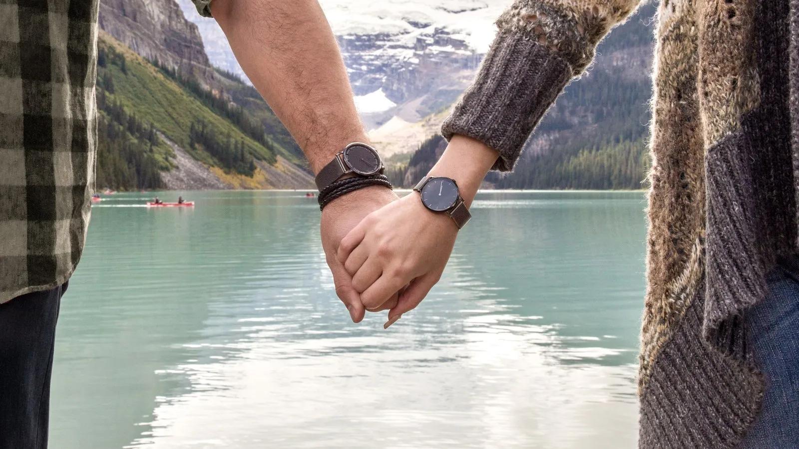 A close-up of a couple holding hands and wearing dark watches, set against a backdrop of a scenic turquoise mountain lake.