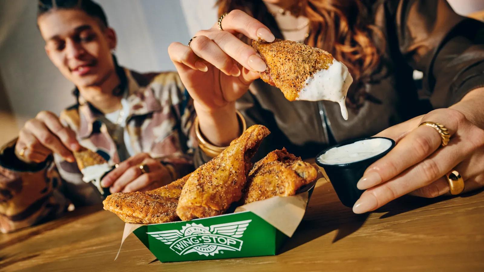 A person dipping a Wingstop chicken wing into ranch sauce with a paper tray of wings on the table.