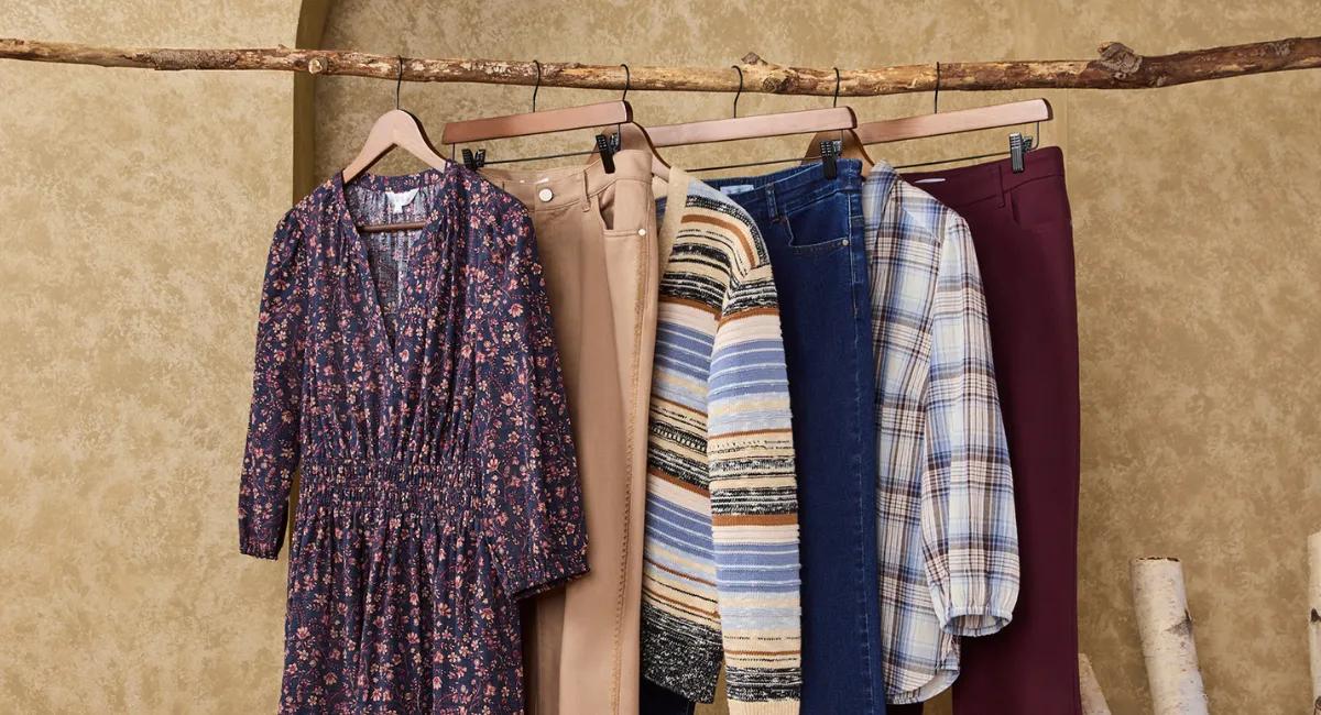 An assortment of fall clothing, including a floral dress, sweaters, jeans, and trousers, hanging on a wooden branch rack.