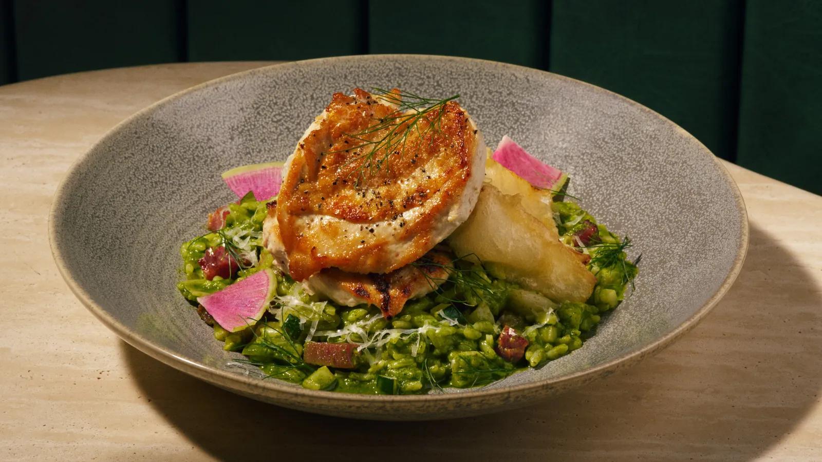A seared chicken breast served over green risotto and garnished with pink radish slices in a speckled gray bowl.