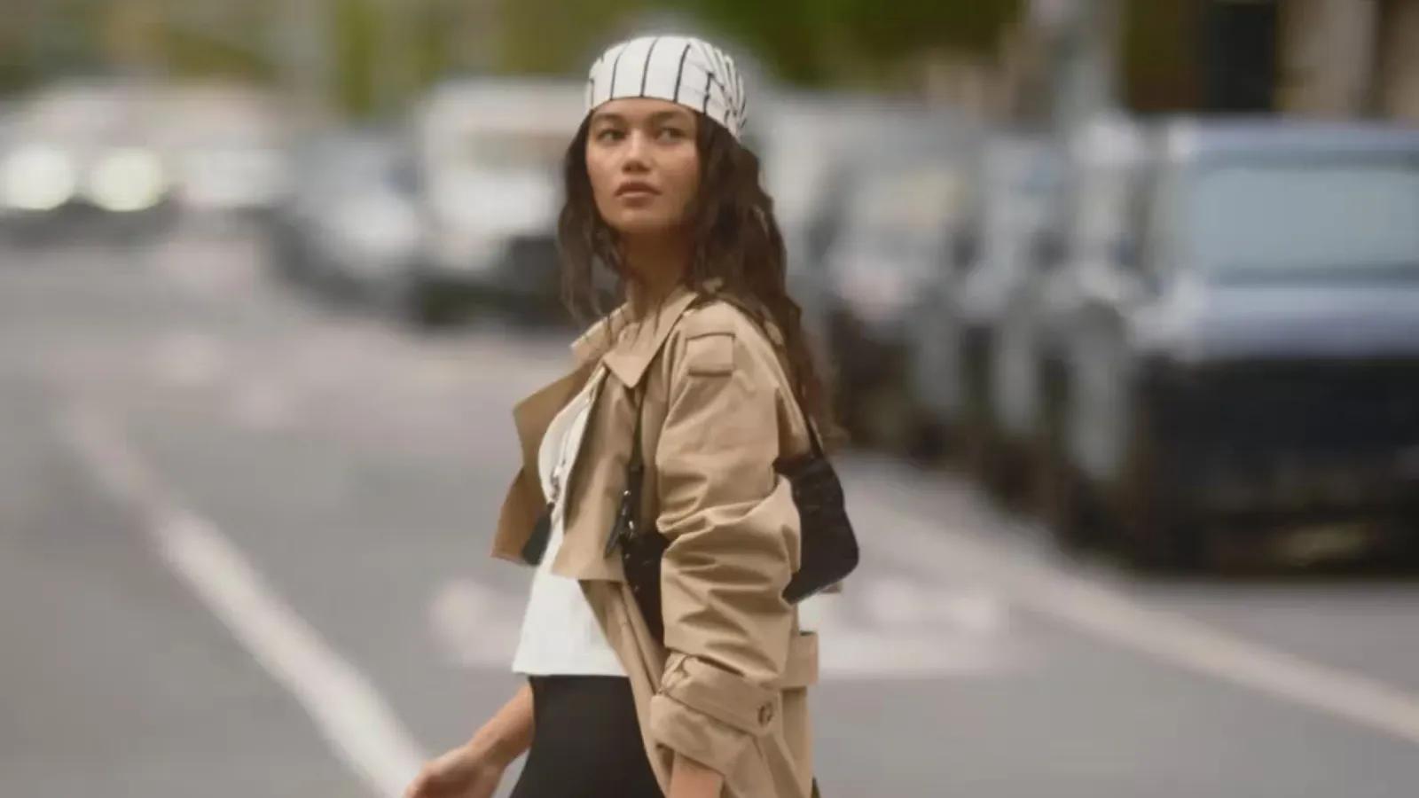 A woman wearing a beige trench coat, a striped headscarf, and a black shoulder bag crossing a city street.