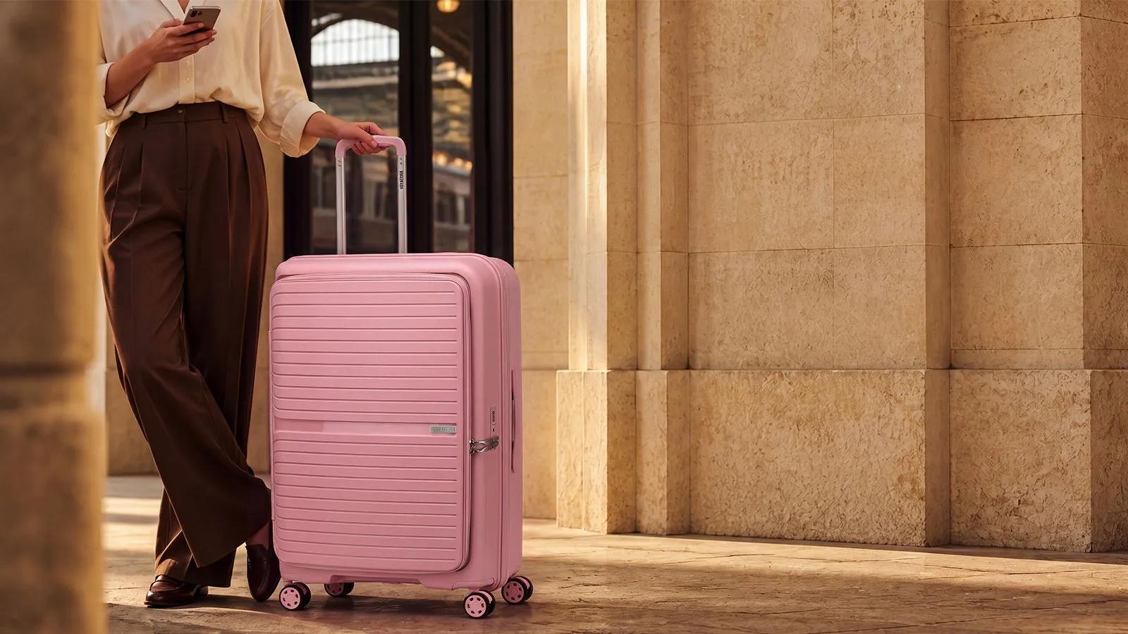 A traveler standing with a pink Voyatique rolling suitcase while using a smartphone in a stone concourse.