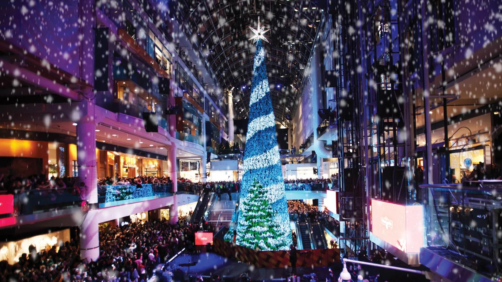 A dazzling holiday display featuring a massive Christmas tree illuminated in blue and white lights, surrounded by crowds inside a mall. Falling snow adds to the festive atmosphere, likely captured during a tree-lighting ceremony or seasonal celebration.