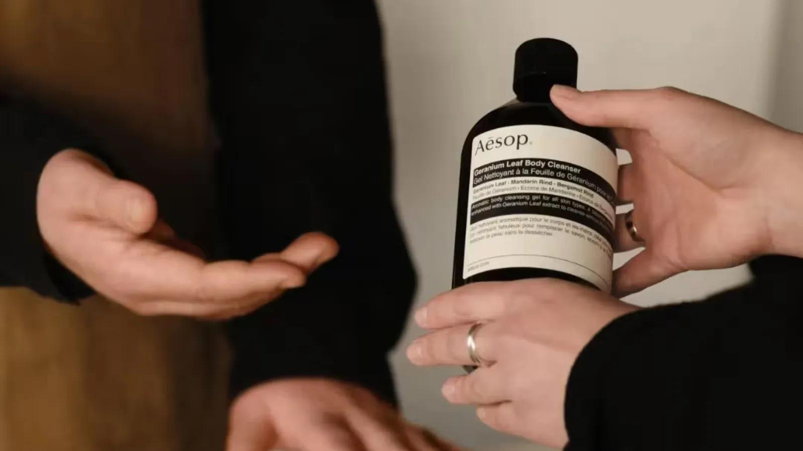 A person holding a bottle of Aēsop Geranium Leaf Body Cleanser while showing it to another person.