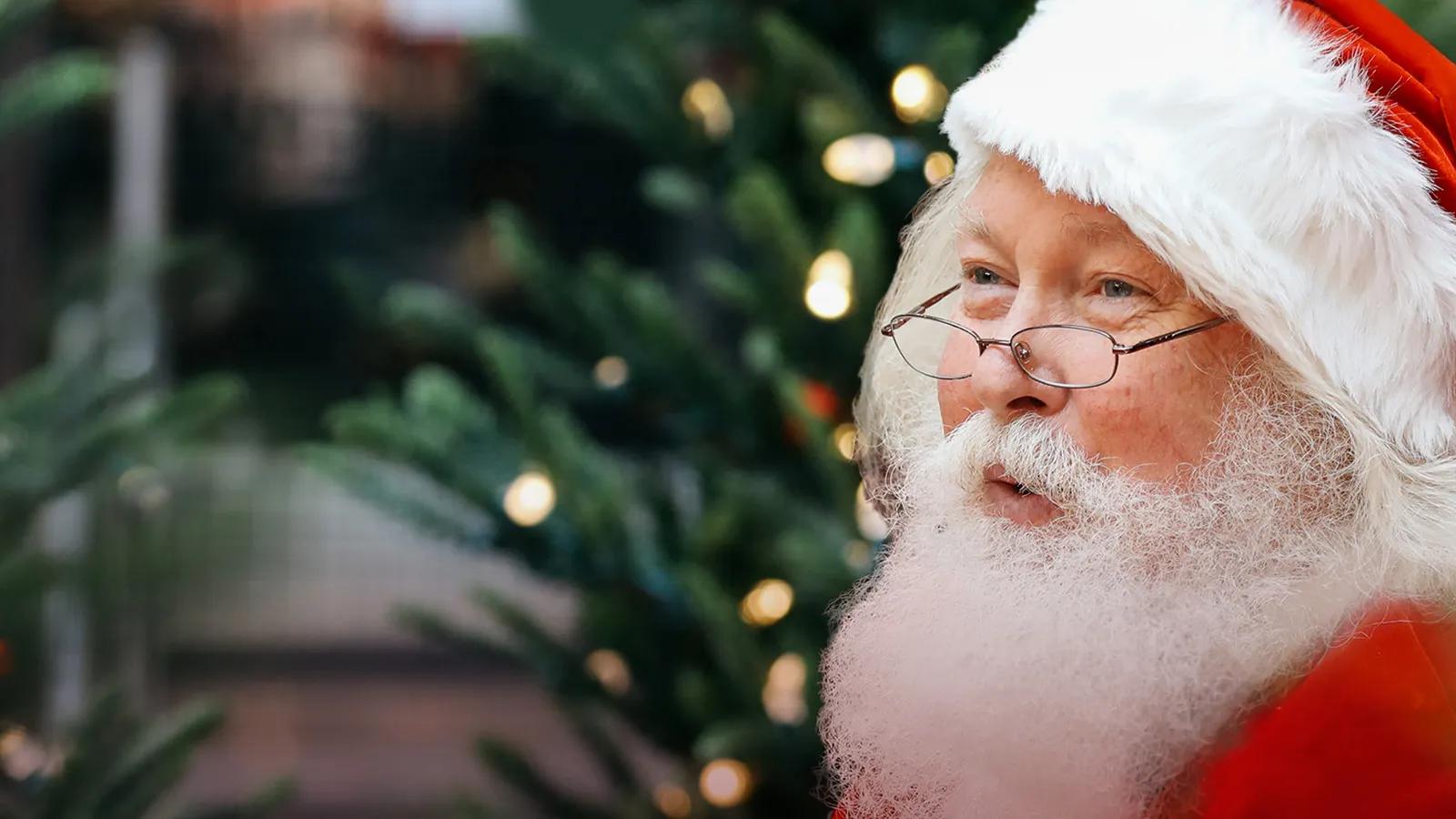 Santa Claus smiling with a Christmas tree in the background.