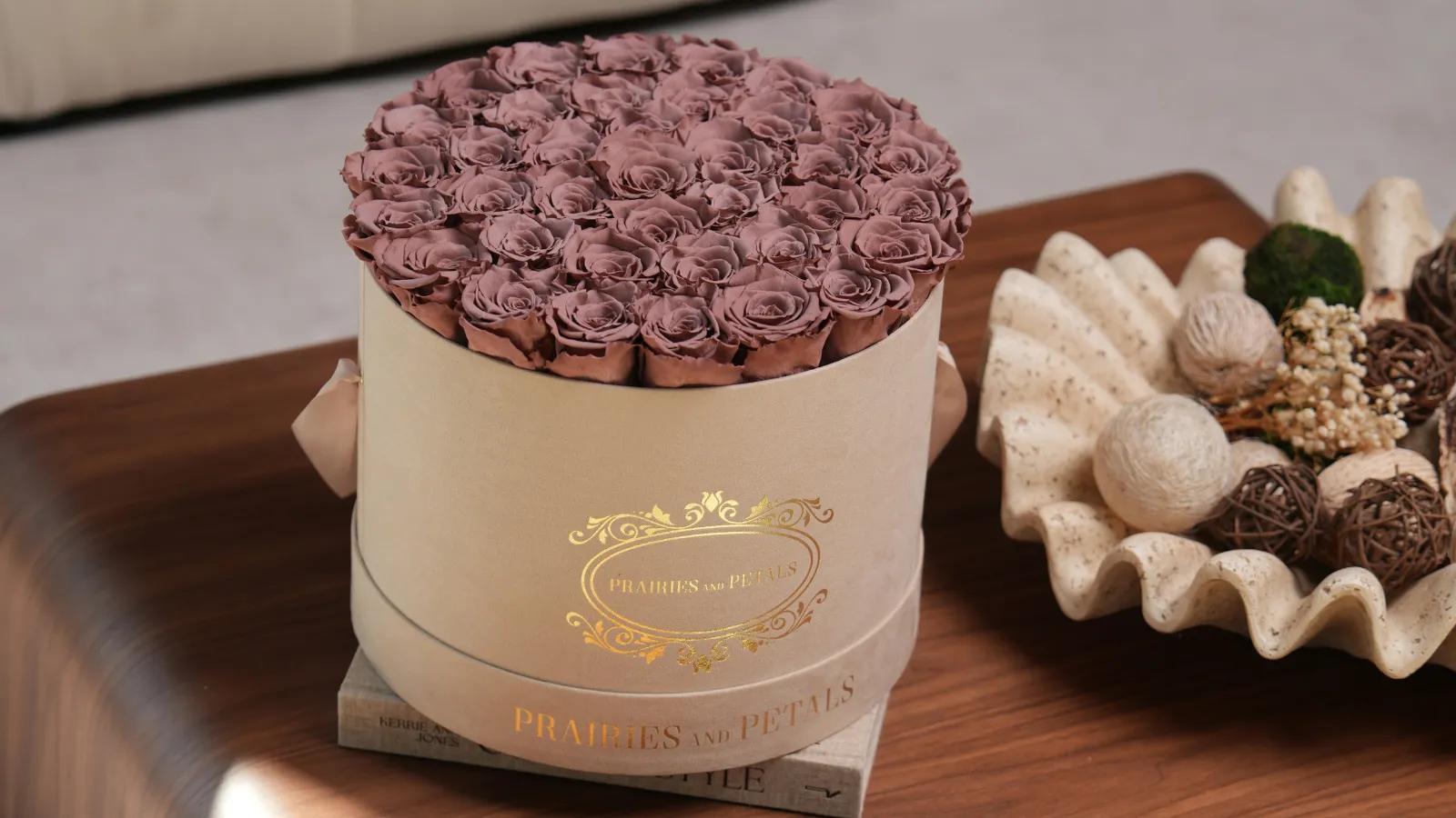 A round beige Prairies and Petals box filled with dusty rose roses resting on a wooden table.