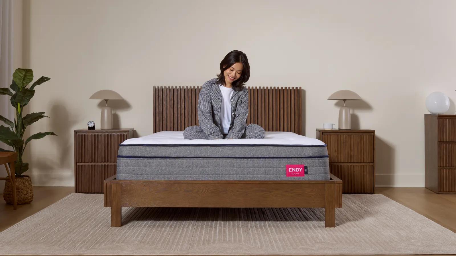 Une femme en pyjama assise les jambes croisées sur un matelas Endy dans un cadre de lit en bois.
