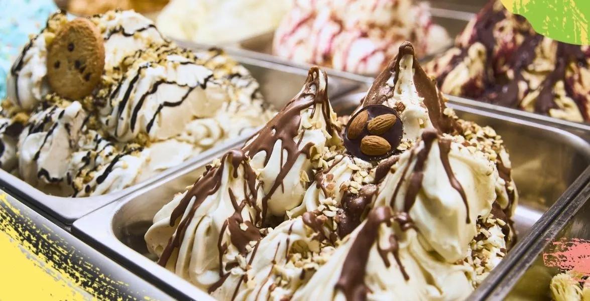 Assorted gelato flavors in metal display pans, topped with chocolate drizzle, nuts, and cookies.