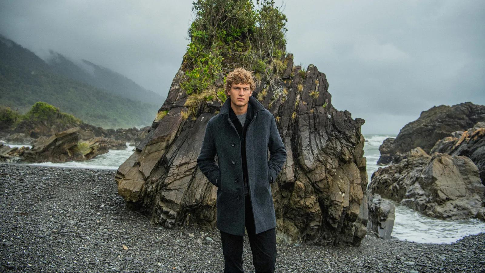 A man wearing a dark Rodd & Gunn wool coat standing on a rocky beach against a moody ocean backdrop.