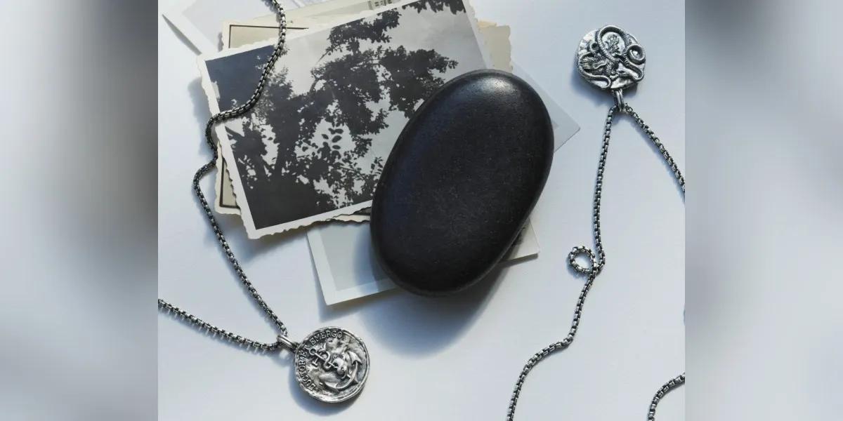 Two silver pendant necklaces arranged with a smooth black stone and black-and-white photos on a white surface.