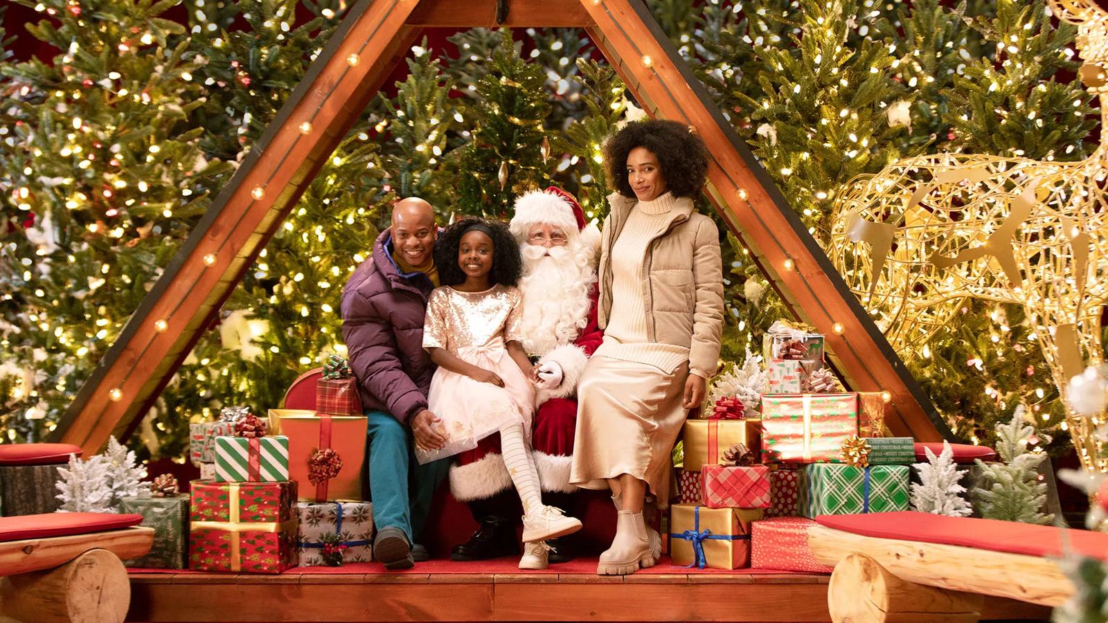 A family of three poses with Santa Claus in a festive Christmas setting. The parents and their young daughter are seated on a wooden bench. Santa sits at the center, dressed in his traditional red and white suit. The scene evokes the warmth of the holidays