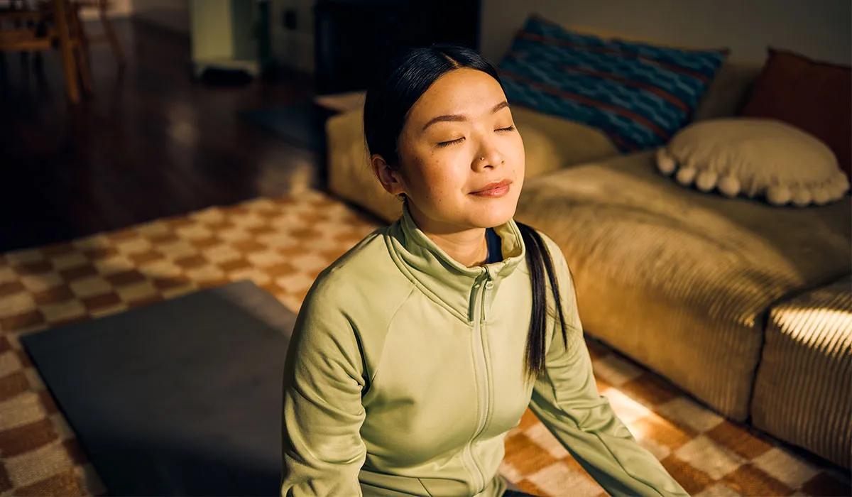 A person in a green jacket sits cross-legged on a mat in a living room, eyes closed, appearing calm.