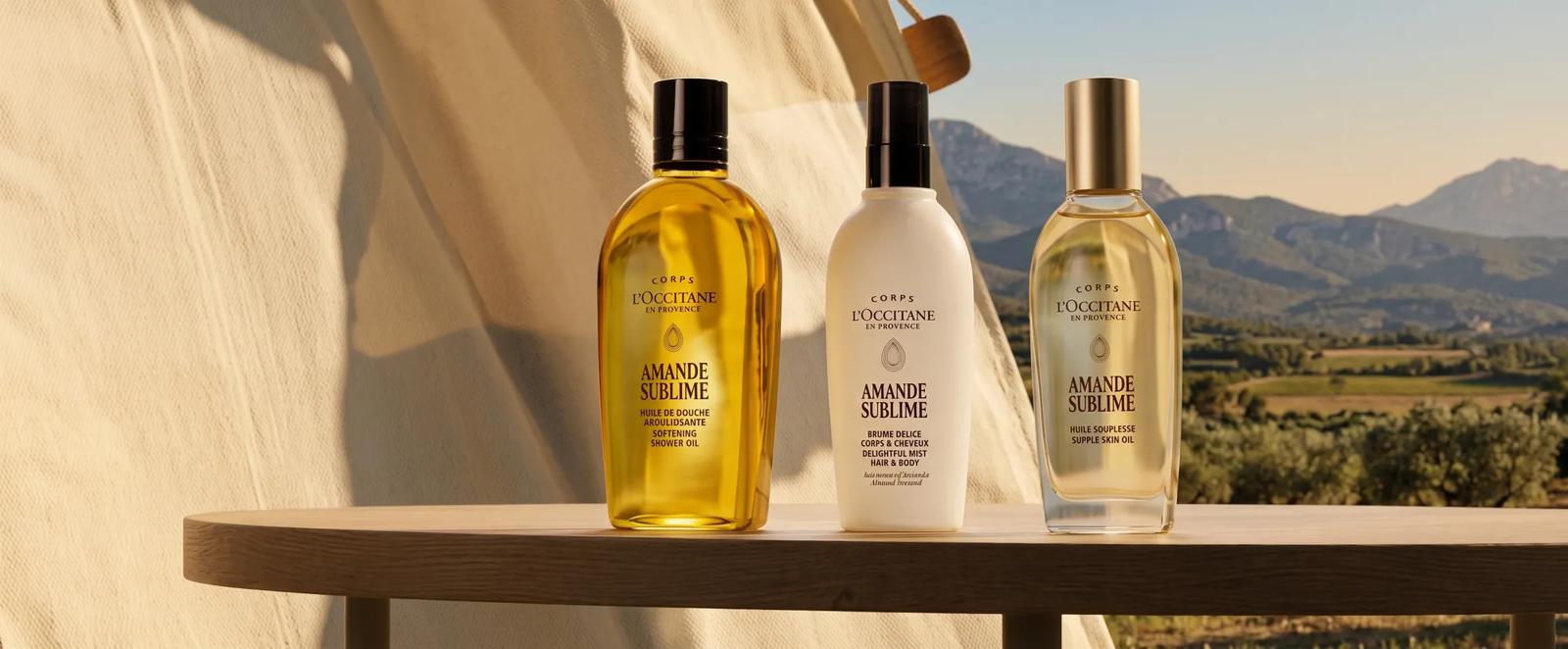 Trois produits pour le corps et les cheveux L'Occitane Amande Sublime exposés sur une table à l'extérieur avec des montagnes en arrière-plan flou.